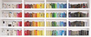 a white bookshelf with 16 shelves. The books are arranged in a rainbow by color going from left to right.