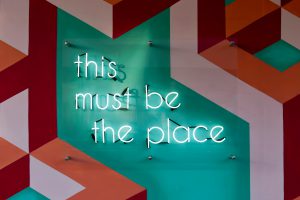 neon lights spell out "this must be the place". Background is geometric art. Art is red, aqua, and a dusty pink.