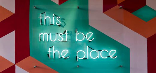 neon lights spell out "this must be the place". Background is geometric art. Art is red, aqua, and a dusty pink.