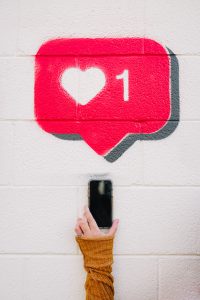 Street art on brick. Art is of a hand with white skin holding a phone. The hand has a silver ring on the ring finger and is wearing a dark yellow sweater. Over the phone is a red message box with a heart and the number 1.