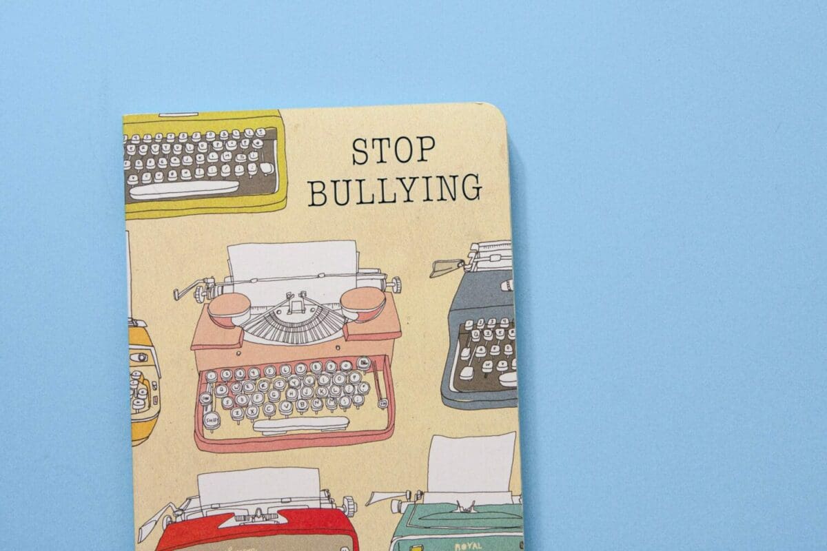 A blue background; an image of a tan writing journal with images of typewriters on the cover in various colors; the cover of the journal has the words "STOP BULLYING"