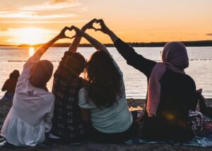 Four people sitting on shore forming two hearts with their hands during golden hour