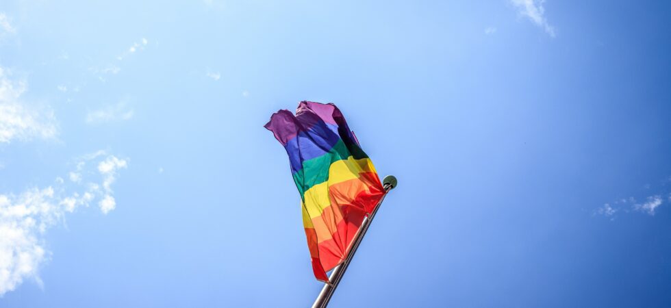 This is a picture of a blue sky with a flag; the picture was taken at an angle at the base of the flag. The flag is a rainbow flag.