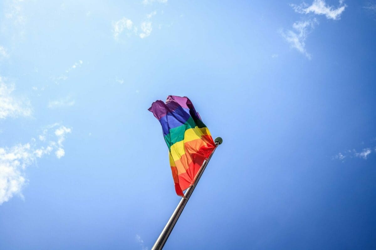 This is a picture of a blue sky with a flag; the picture was taken at an angle at the base of the flag. The flag is a rainbow flag.