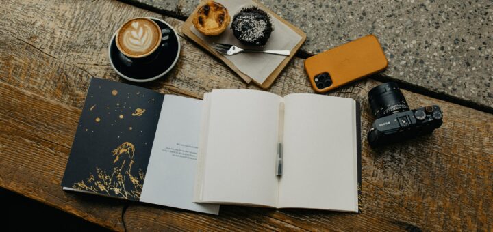 An open journal sitting on top of a wooden table next to a cup of coffee, a phone, a camera, and a plate of deserts