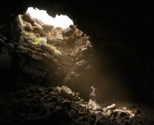 This is a picture of a person in a cave and the person is looking downward and there is a beam of light coming down from an opening at the top of the cave.