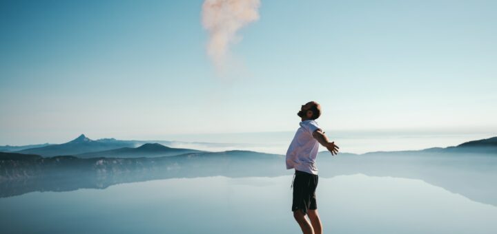 this is a photo of a man clearly in focus standing with arms out stretched looking toward the sky and he is in front of a lake and mountains are in the distance.