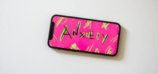 A cell phone lies on a flat white surface. The screen has stylized art reading "Anxiety" Background of the art is hot pink, and the text is black with neon yellow highlights