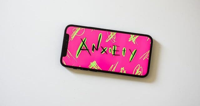 A cell phone lies on a flat white surface. The screen has stylized art reading "Anxiety" Background of the art is hot pink, and the text is black with neon yellow highlights